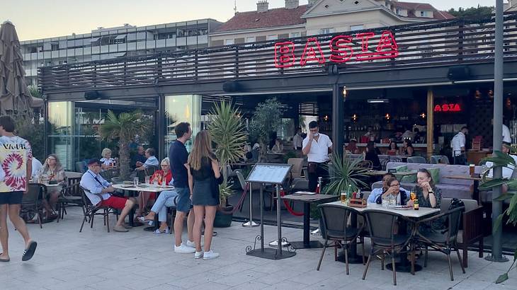 People sitting and walking in front of a restaurant with a red sign 'Basta'