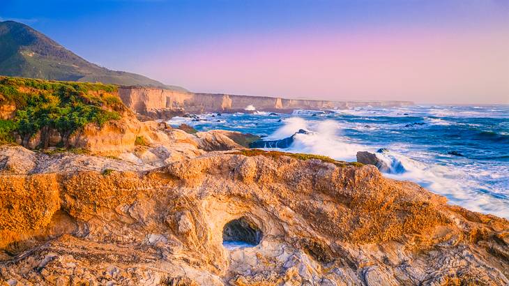 Orange-colored rocks surrounded by crashing waves under a pink and purple sky