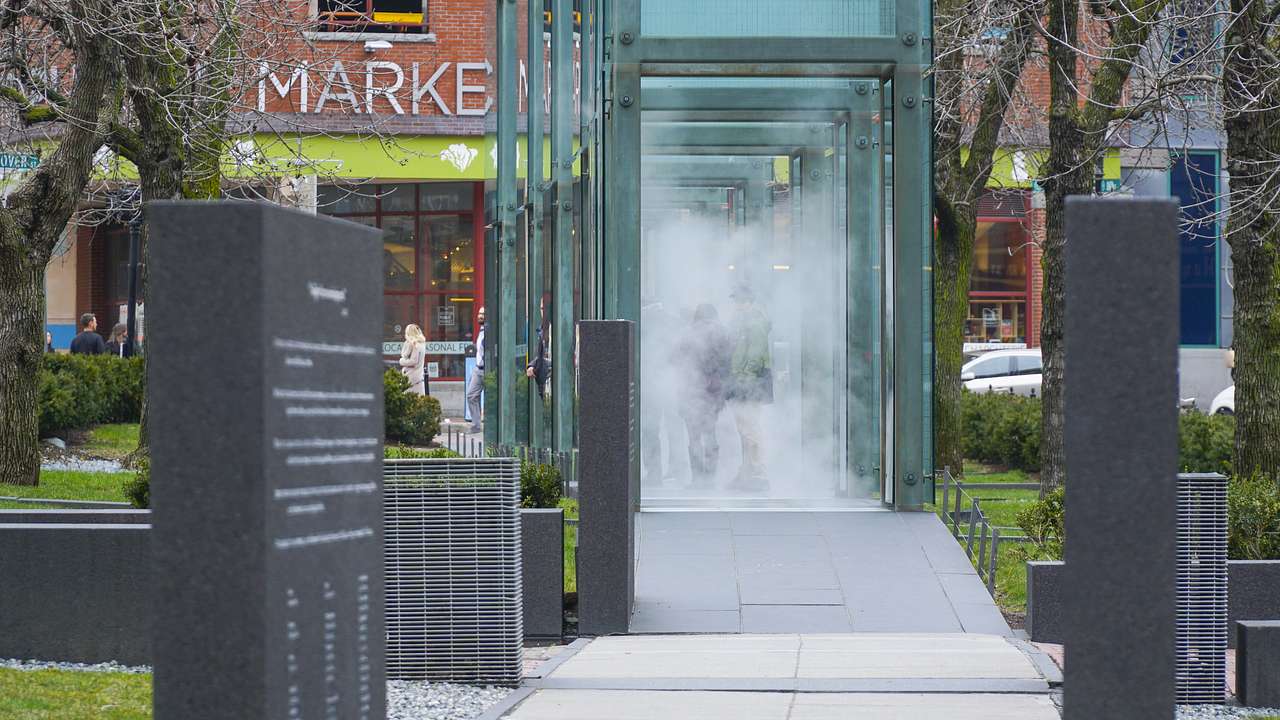 Surrounded by a lawn is a granite pathway with tall glass towers filled with mist