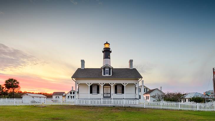 A small house with a lighthouse behind it, grass in front, and a sunset sky