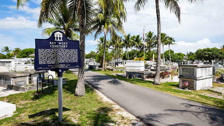A Key West Cemetery sign with gravestones, palm trees, and a path behind it