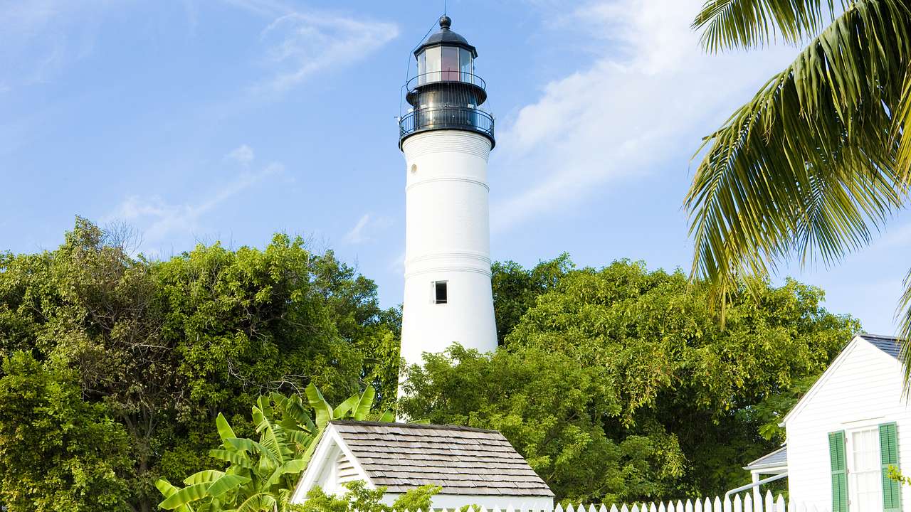 A white lighthouse with black top surrounded by trees with a house and fence in front