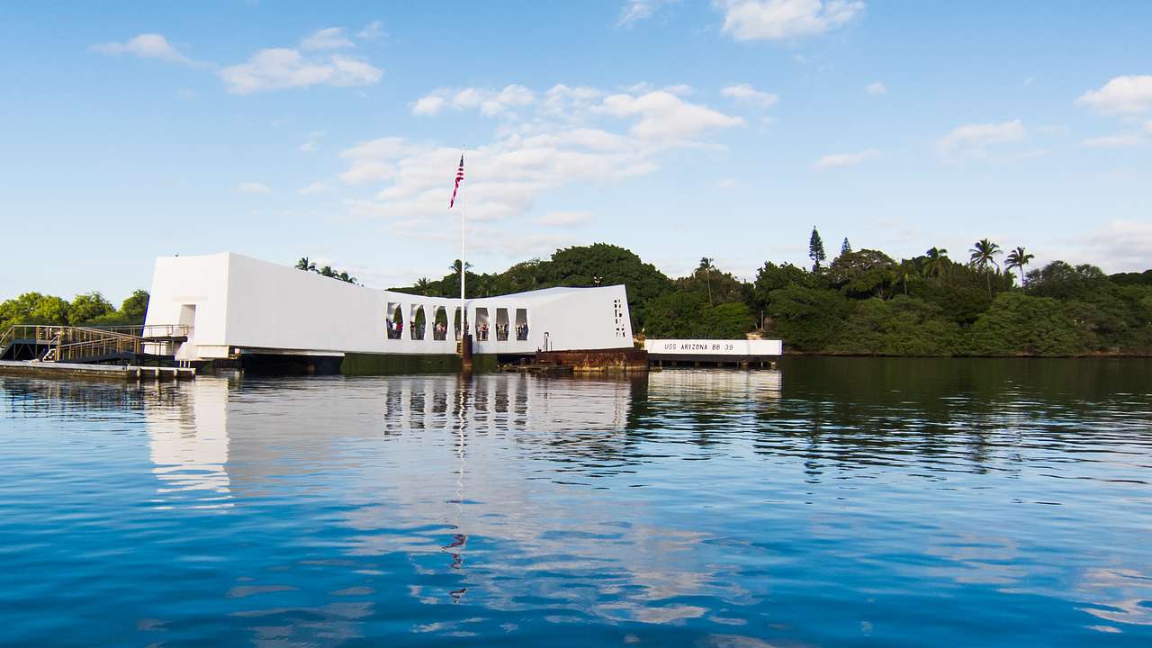 A white memorial building with an American flag and water in front and trees behind