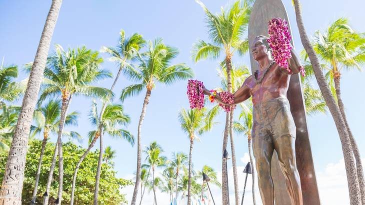 A bronze statue with pink flower leis on it and palm trees behind it