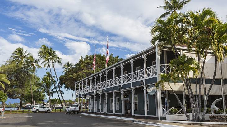 A building with a white balcony, palm trees surrounding it, and two cars in front