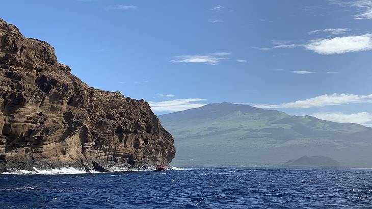 A rocky cliff with a mountain in the background and ocean water surrounding it