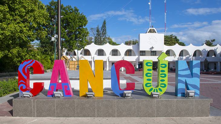 A 3D colorful Cancun sign with a building and trees behind it