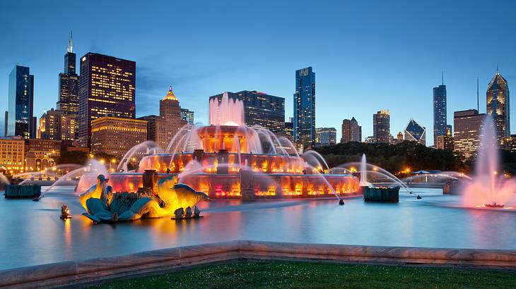 An illuminated water fountain at night with skyscrapers behind it