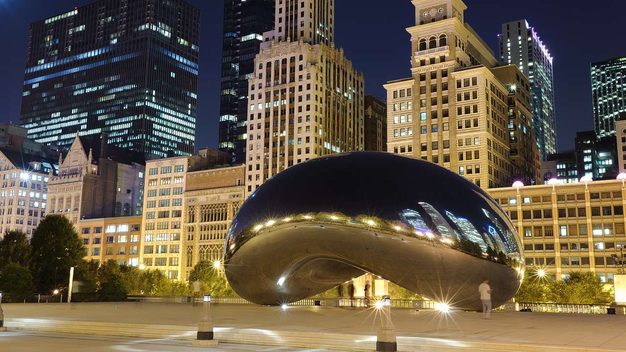A bean-shaped reflective sculpture with skyscrapers behind it at night