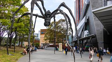 A large spider sculpture on a path walk surrounded by buildings, trees and people
