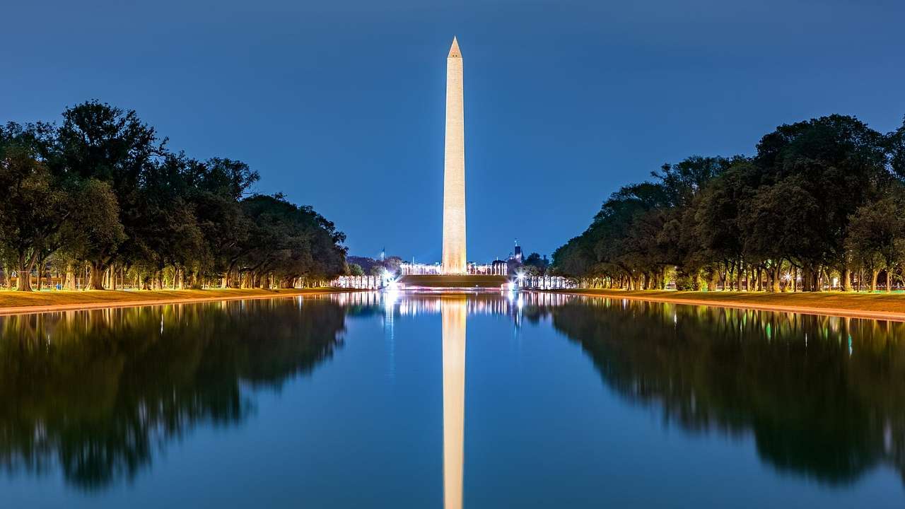 A white obelisk reflecting on the water at night with trees surrounding