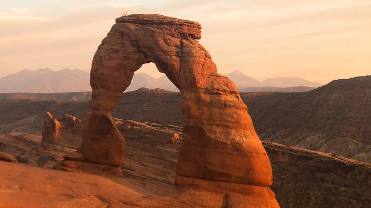 A uniquely-shaped sandstone arch surrounded by canyons at sunset