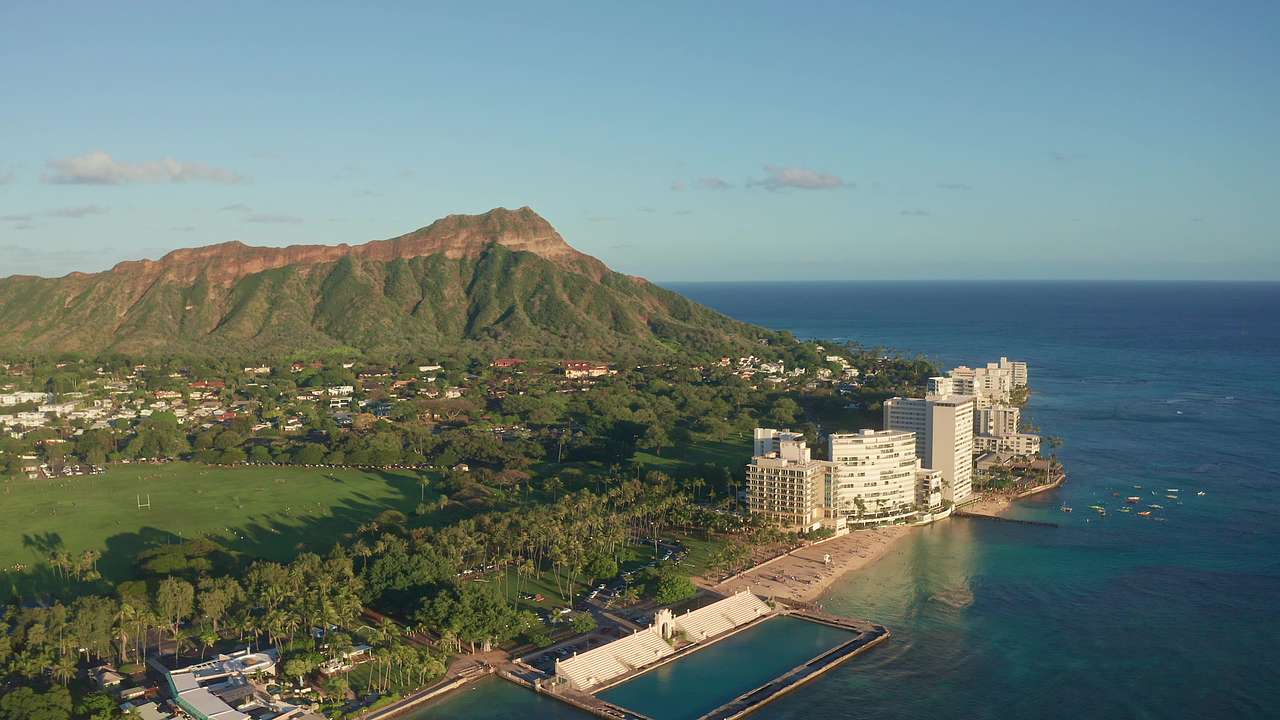 A large greenery-covered mountain with buildings to the side, surrounded by ocean