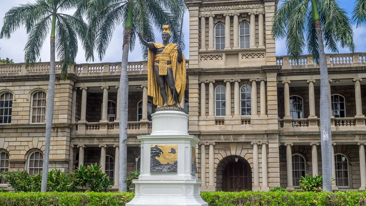 A gold statue on the grass in front of a palace building with palm trees surrounding
