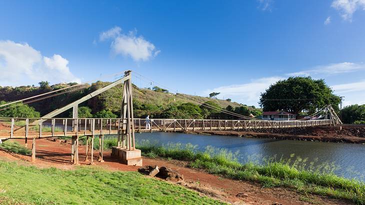 A wooden suspension bridge over a river with a mud path and grass to one side