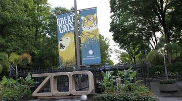 A concrete zoo sign with a banner above it with big cats on and greenery surrounding