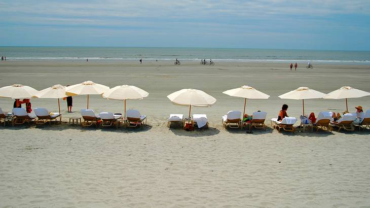 People relaxing on beach chairs with white umbrellas and biking along the shore