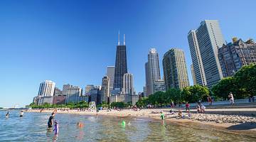 An urban skyline with a sandy beach and people in the water in front of it