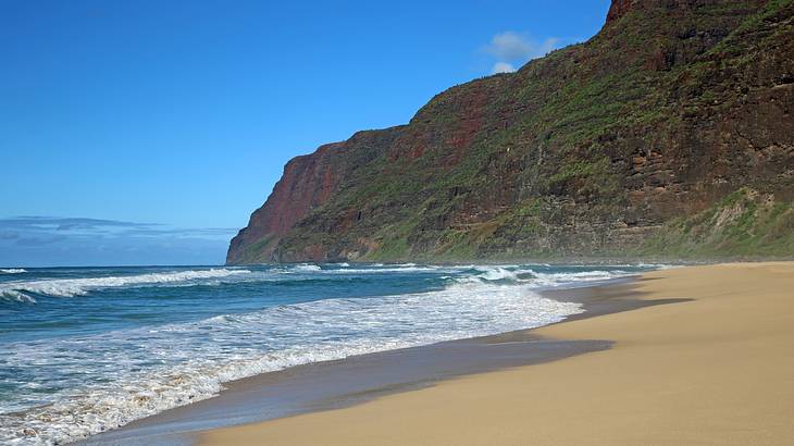 Sand with waves crashing onto it and a cliff to the side under a blue sky