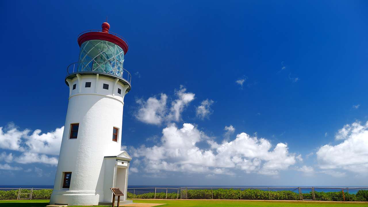 A white lighthouse with a red top sitting on the grass under a blue sky with clouds