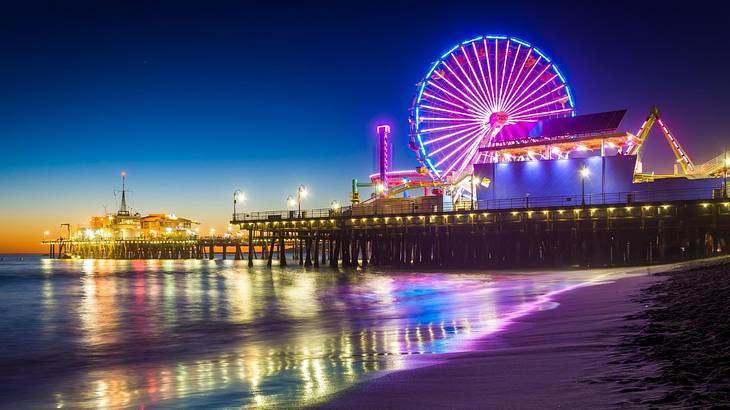 Neon lights of a Ferris wheel and rides reflected on the water by the shore at night