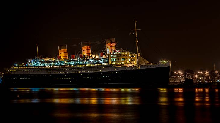 Lights of a docked ship with three funnels reflected in the water at night