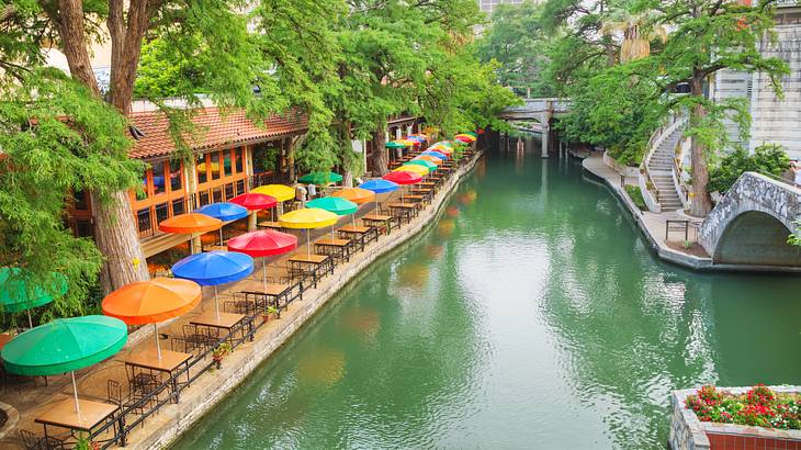 A river with a bridge on one side and trees and colorful patio umbrellas on the other