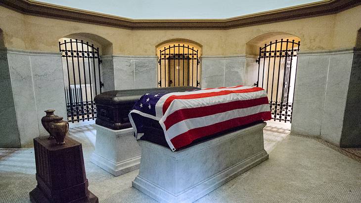 Two caskets, one covered in a US flag, in a stone room with iron gates