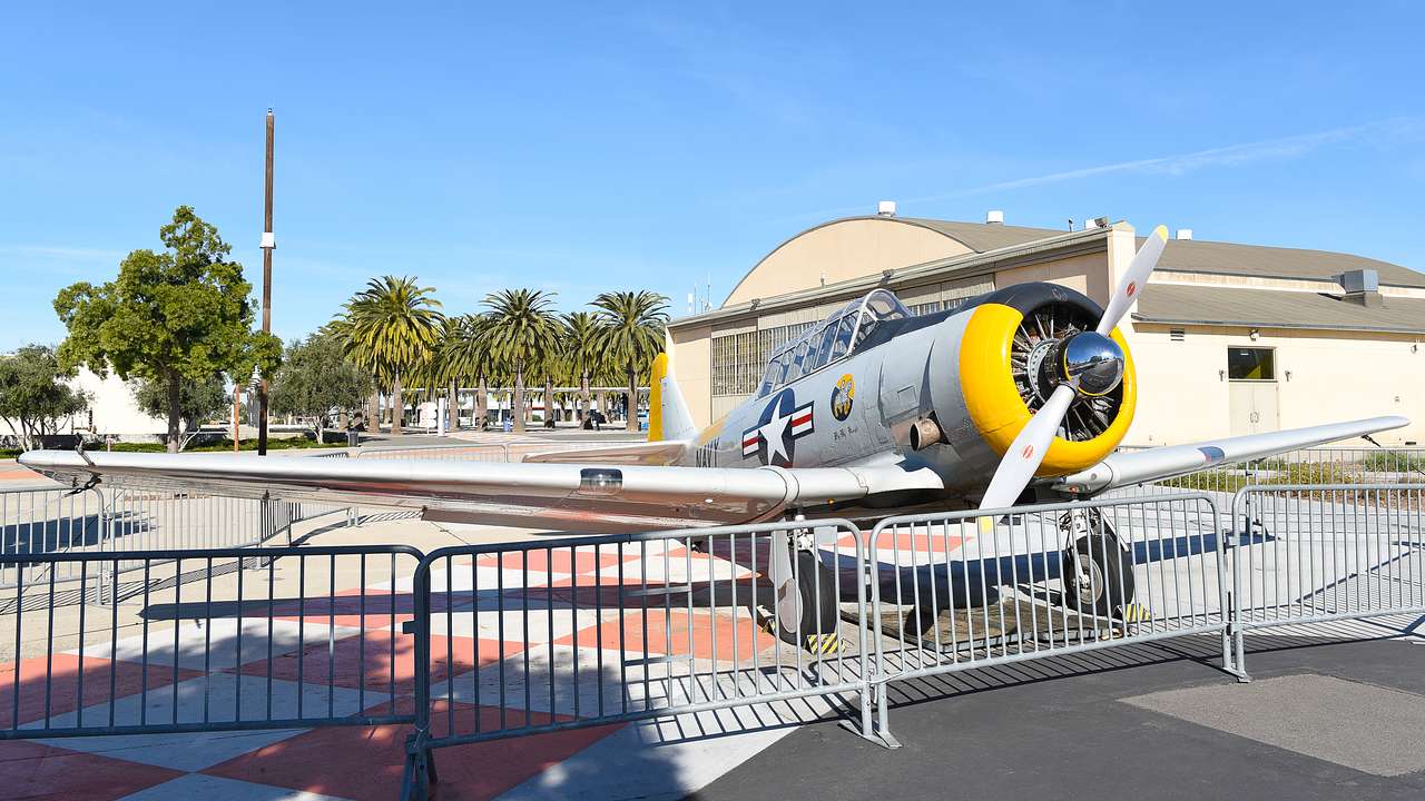 Yellow and gray vintage plane parked in a fenced ground with trees behind