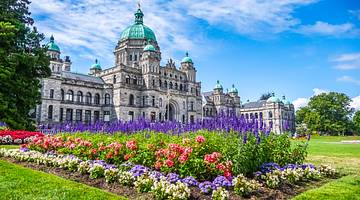 A large building with green domes next to trees facing a garden of colorful flowers