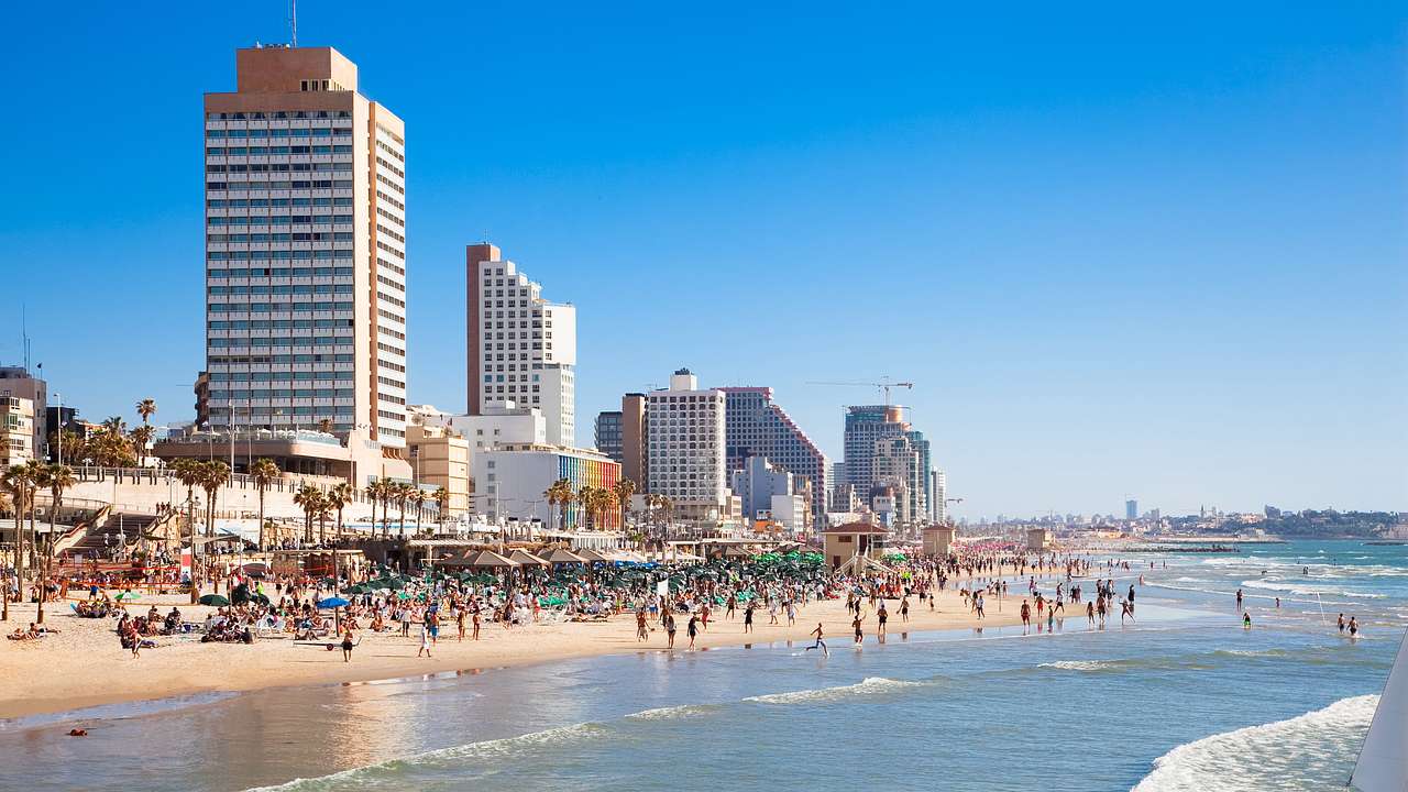 A crowded beach with people on the sand and in the water with tall buildings behind