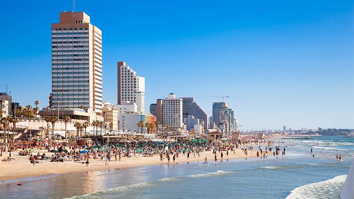 A crowded beach with people on the sand and in the water with tall buildings behind