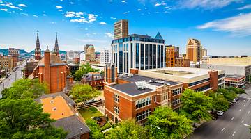 A city skyline filled with historic-looking buildings, greenery, a road, and blue sky