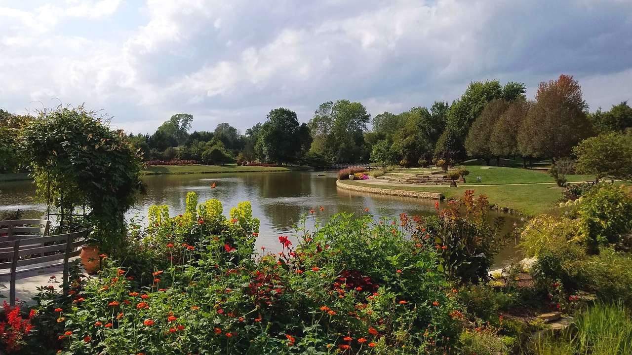 A lush green garden with green bushes and red flowers surrounding a water body