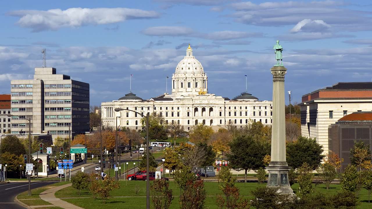 A white state capitol building with a dome roof and a tall monument statue and park