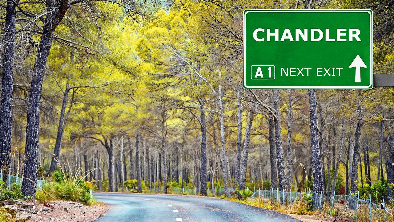 A green road sign with "Chandler" and other text written on it above a forest road