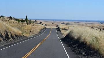 A grey paved road running through a dry grassy landscape on a clear blue day