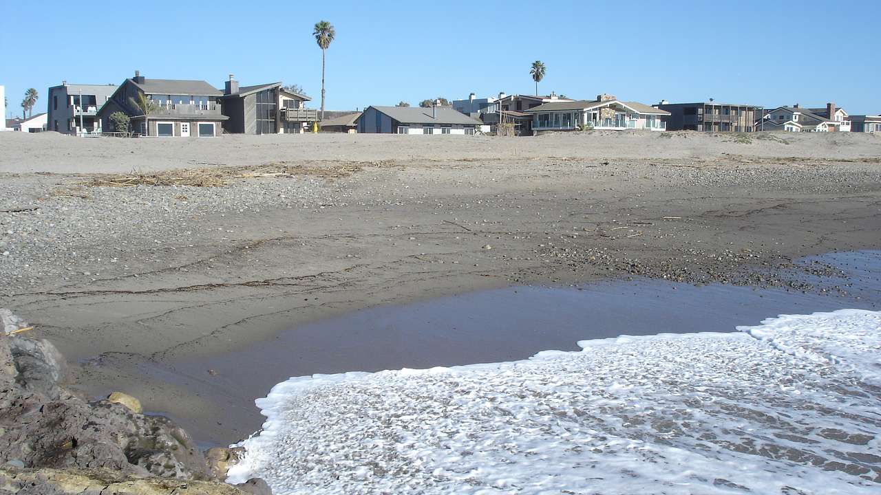 A grey sandy beach with grey houses at the back and rocks on the bottom left