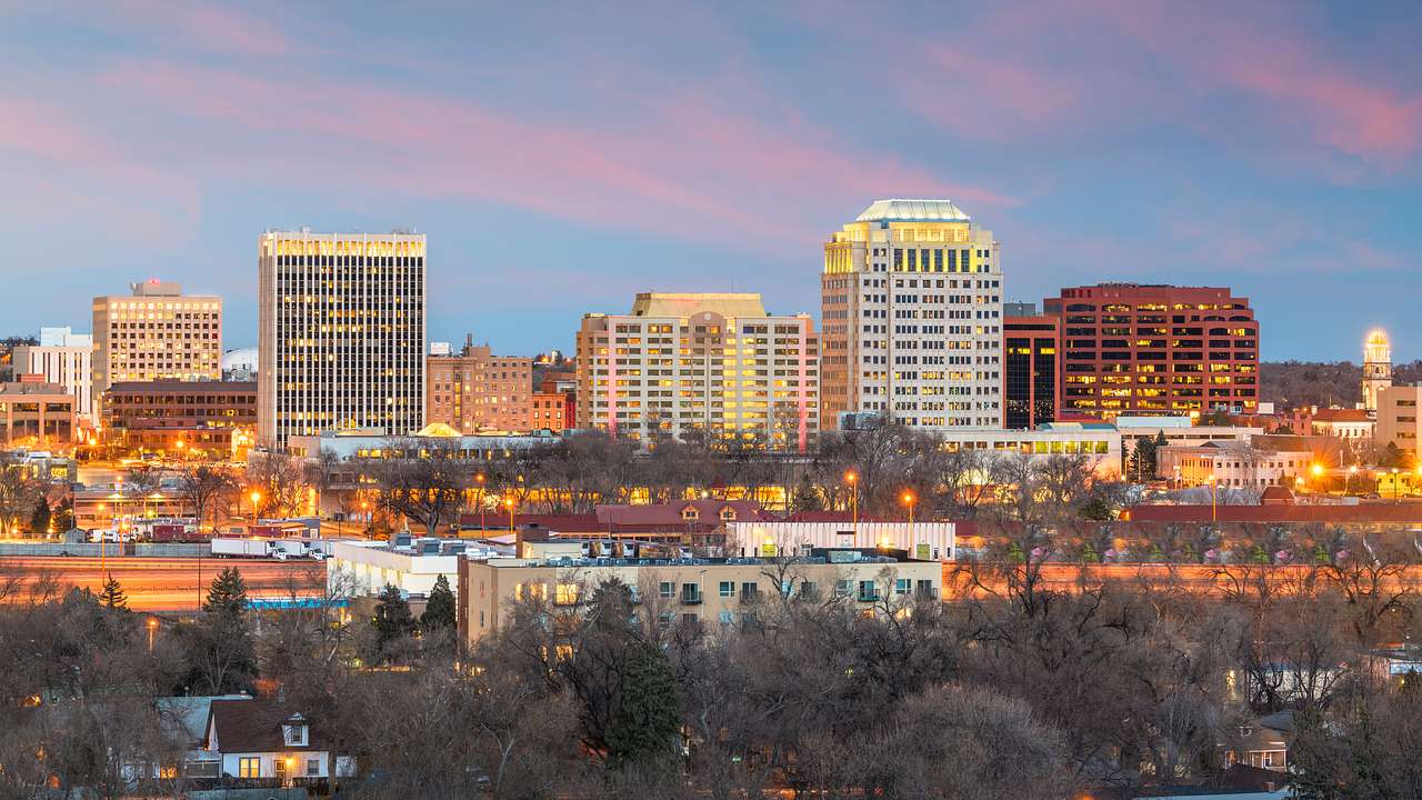 A city skyline with concrete buildings at the back and trees in front at sunset