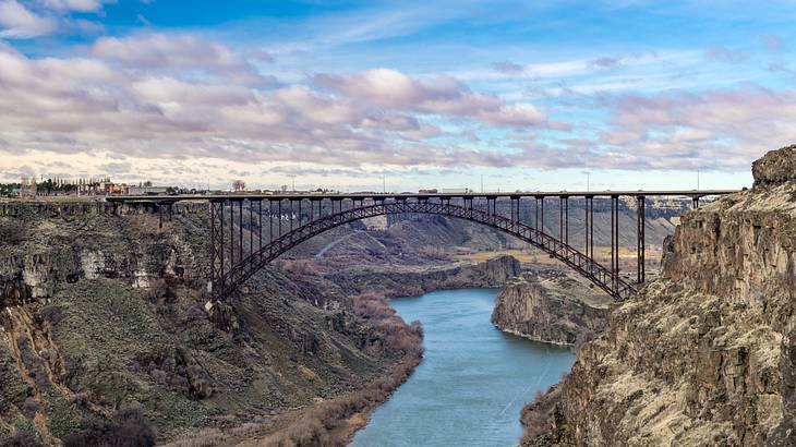 A side view of an arched bridge with cars over a river connecting two cliffs