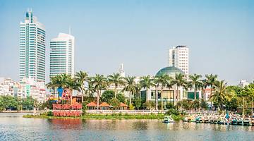 A skyline view of 3 tall buildings behind a row of palm trees facing a body of water