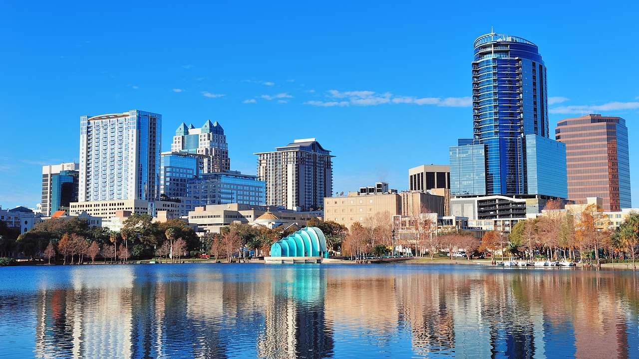 Blue, sunny skyline of buildings with brown trees at the base, facing a body of water