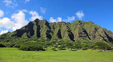 Mountain with vertical ridges and trees and green fields at its base on a sunny day