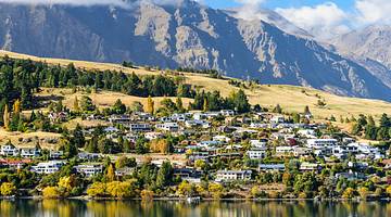 Buildings on a hilly incline with trees facing a body of water, with mountains behind