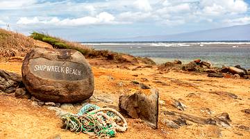 Sandy, rocky beach, and a "Shipwreck Beach" sign on a rock, under a partly cloudy sky