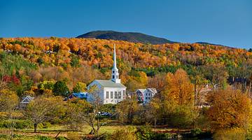 A small white church with a pointy top, among hills, in the middle of fall foilage