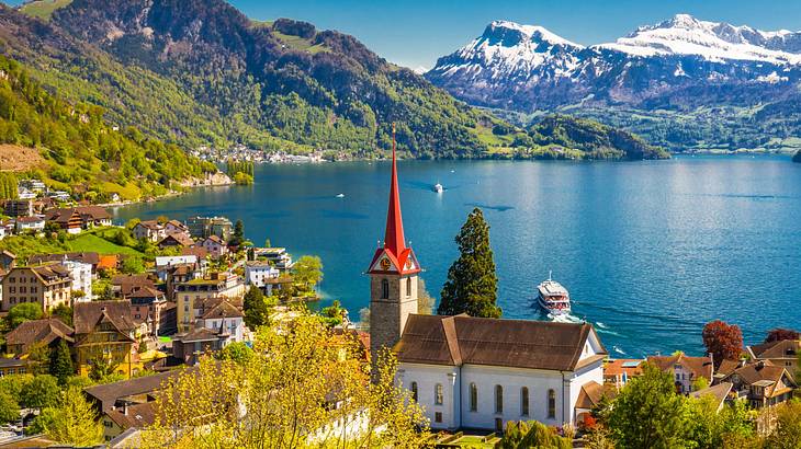 A church with a red spire on the roof with a lake and snow-capped mountains behind