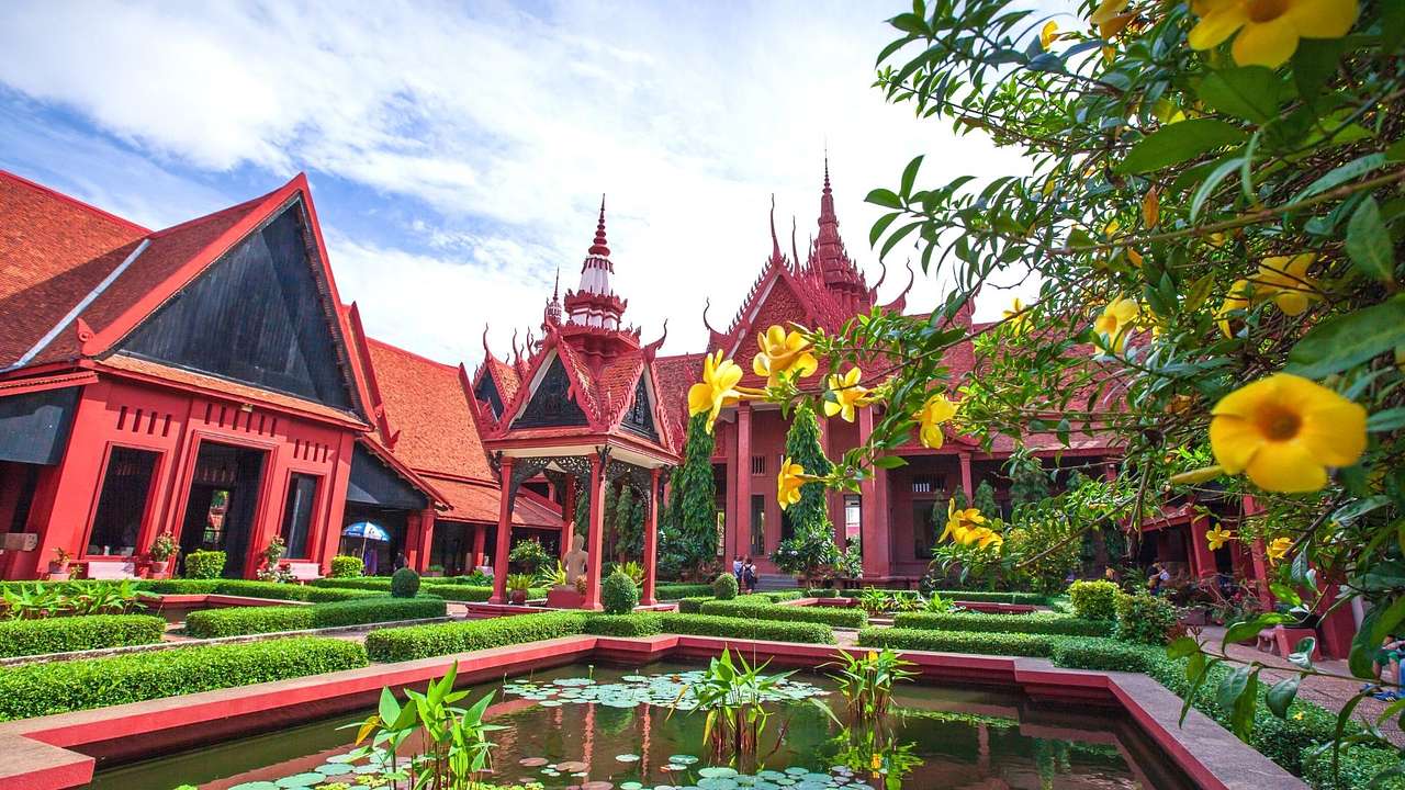 A red-lined pond surrounded by red Khmer architecture and green and yellow plants