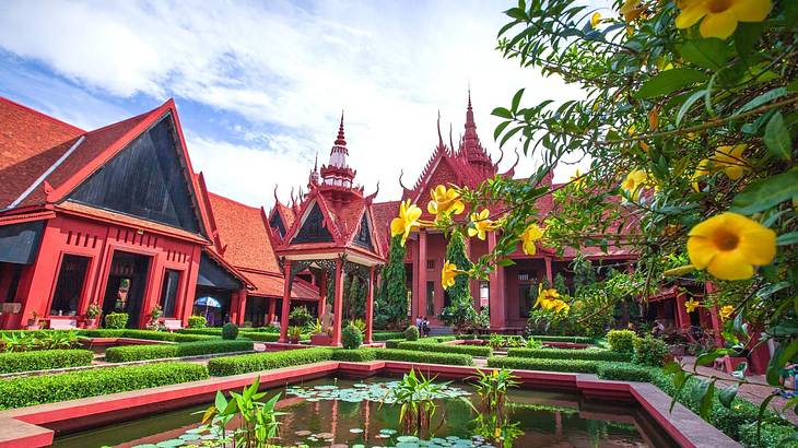 A red-lined pond surrounded by red Khmer architecture and green and yellow plants