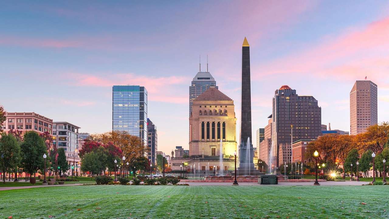 A city skyline with buildings and a tall thin memorial and green lawn in front